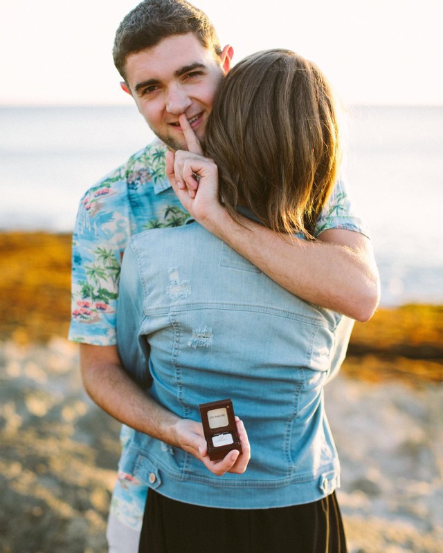 couple hugging while man holds engagement ring behind woman's back